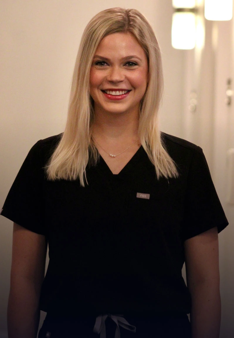 Woman smiling at camera in black scrubs inside dental office hallway.