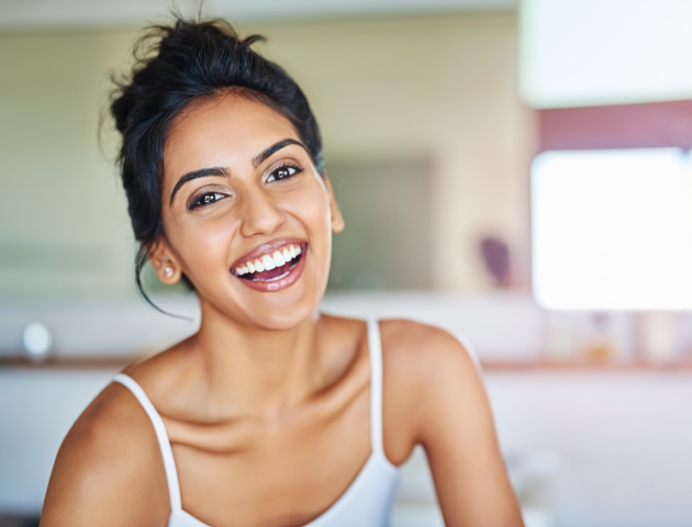 Smiling woman in bright kitchen setting, wearing white tank top and hoop earrings.