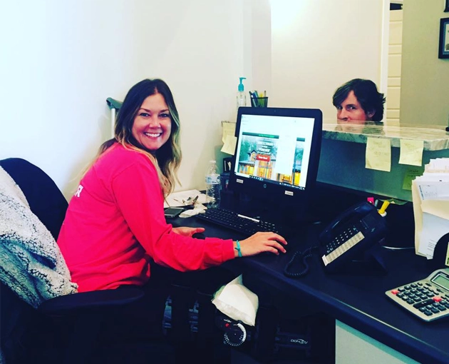 Two women smiling at computer workstations in a dental office setting.