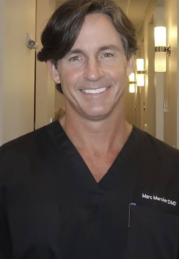 Male dental professional in black scrubs smiling at camera in clinical hallway setting.