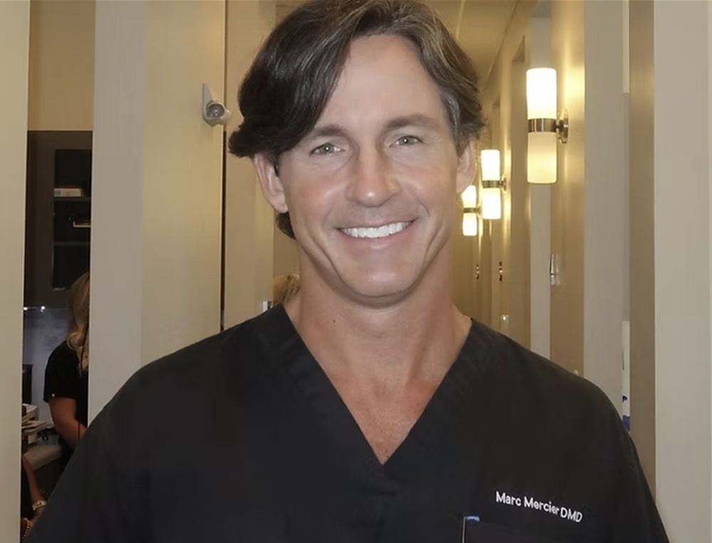 Male dental professional smiling at camera wearing black scrubs in modern dental office hallway.