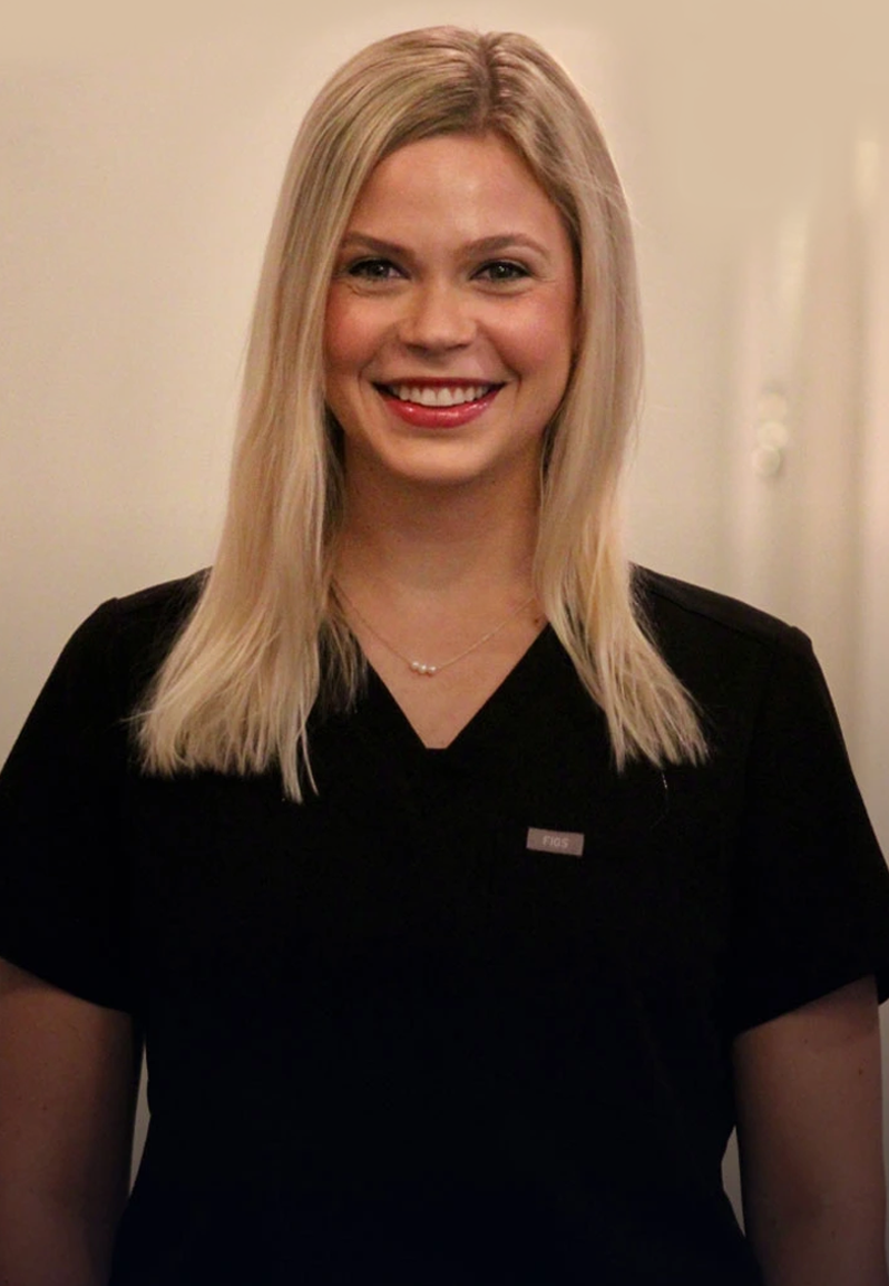 Professional woman with blonde hair smiling at camera in clinical dental office setting, wearing black scrubs.